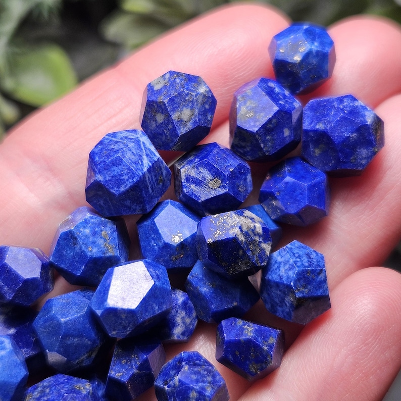 May include: A collection of small, faceted lapis lazuli gemstones. Each stone features a deep blue color with natural golden pyrite inclusions and a multi-sided geometric cut, creating a reflective surface across the various individual pieces.