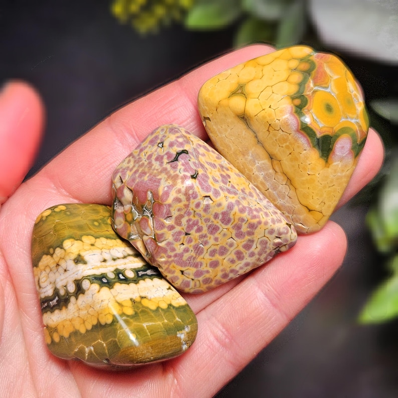 May include: Three polished gemstones in shades of yellow, green, and purple are held in a hand. The stones have unique patterns and textures. The gemstones are likely used for decorative purposes or in jewelry.