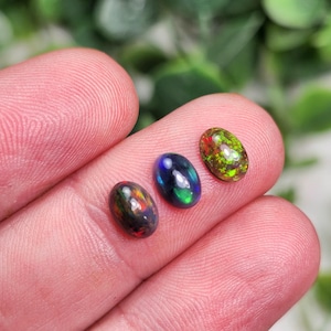 May include: Three oval-shaped opals in shades of black, blue, green, red, and yellow. The gemstones are displayed against a blurred green background. The opals are of varying sizes and exhibit a play of color.