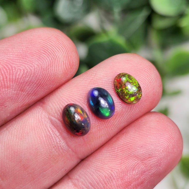 May include: Three oval-shaped opals in shades of black, blue, green, red, and yellow. The gemstones are displayed against a blurred green background. The opals are of varying sizes and exhibit a play of color.