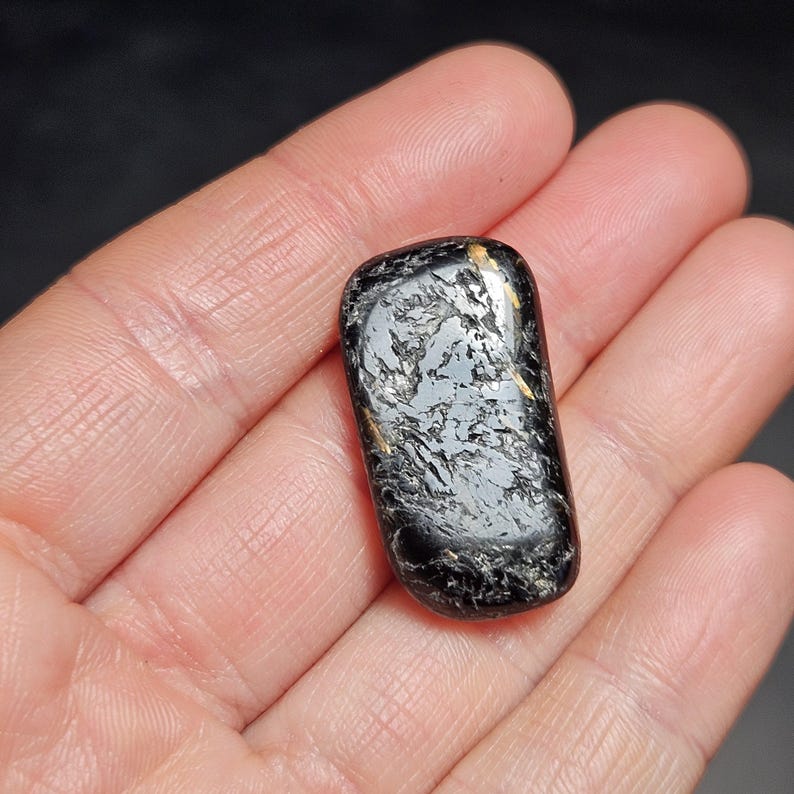 May include: A polished, rectangular black stone with a marbled, silver-gray pattern and gold flecks. The stone is held in an open hand, showcasing its smooth, rounded edges. This gemstone is likely used for jewelry or decorative purposes.