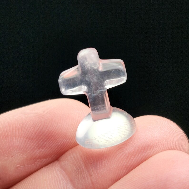 May include: A small, clear, cross-shaped object with a rounded base. The cross is transparent and appears to be made of a smooth, possibly glass or crystal-like material. The object is held against a dark background.