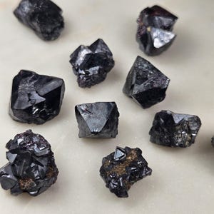 Puede incluir: Una colecci&oacute;n de cristales oscuros y facetados de color negro intenso. Los cristales var&iacute;an en tama&ntilde;o y forma, con bordes afilados y una superficie reflectante. La disposici&oacute;n sugiere una exhibici&oacute;n de gemas naturales en bruto, posiblemente con fines metaf&iacute;sicos o decorativos.