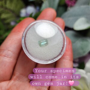 May include: A small, light blue and green gemstone in a clear plastic container with white sand. The text "Your specimen will come in its own gem jar!" is below the container.