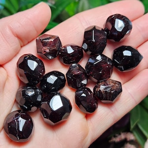 May include: A collection of 14 dark red garnet tumbled stones, each with a faceted, geometric shape. The stones are arranged in a hand, showcasing their unique facets and deep red color.