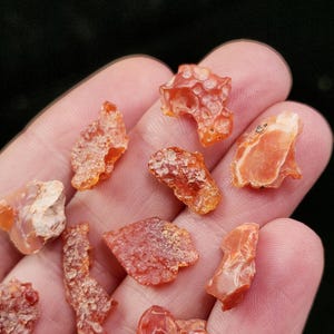 May include: A collection of raw, orange carnelian gemstones of varying shapes and sizes. The stones have a translucent quality, with some displaying a layered appearance. The carnelian pieces are held in a hand against a dark background.