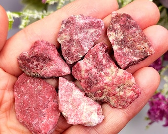 Puede incluir: Una colección de piedras preciosas de turmalina rosa en bruto. Las piedras varían en tamaño y forma, con una gama de tonos rosados que van del claro al rosa intenso. Algunas piezas tienen una mezcla de rosa y blanco. Las piedras preciosas se sostienen en una mano.
