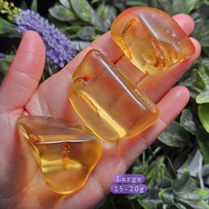 May include: Three polished amber stones with a translucent golden-yellow hue and visible internal organic inclusions. The stones are smooth and irregular in shape. A purple label on the image indicates a large size range of 15-20 grams.