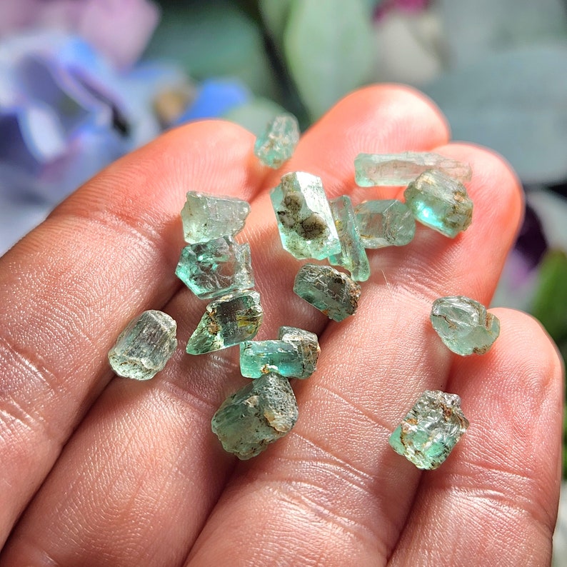 May include: A collection of small, green emerald crystals with a rough, natural finish. The crystals are various shapes and sizes, and some have a slight golden hue.