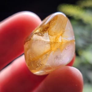 May include: A polished, translucent gemstone with a smooth, rounded shape. The stone features a clear, glass-like appearance with prominent golden-yellow inclusions and internal fractures that create a complex, multi-faceted visual texture.