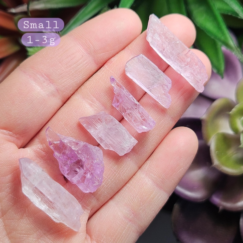 May include: A collection of six small, pale pink to purple selenite crystal points, ranging in size from 1 to 3 grams. The crystals are displayed on a hand, with the text "Small 1-3g" visible in the background.
