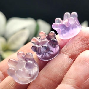 May include: Three translucent, hand-shaped sculptures in varying shades of purple and lavender. The sculptures are held in a hand, with a blurred background. The sculptures have a smooth, polished appearance and are likely made of glass or crystal.