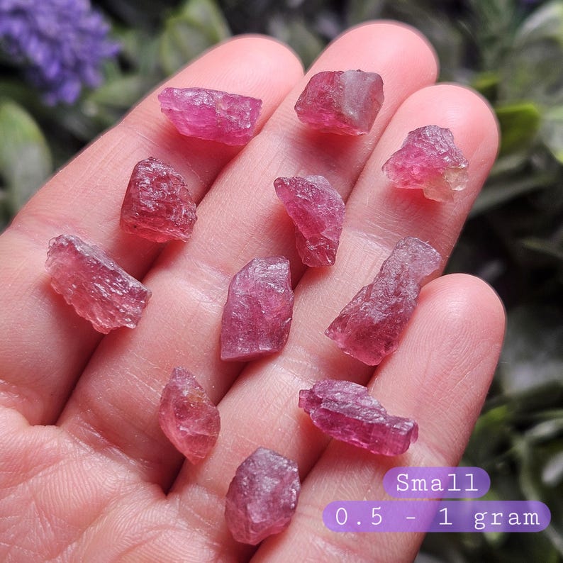 May include: A collection of small, raw, pink tourmaline crystals with irregular, faceted surfaces. The stones vary in shape and size, with a purple text overlay in the bottom right corner stating: Small 0.5 - 1 gram.