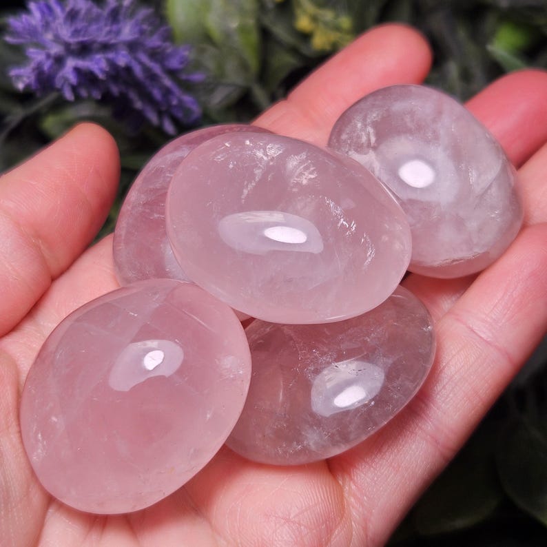K&ouml;nnte beinhalten: Eine Ansammlung glatter, polierter Rosenquarzsteine mit einem durchscheinenden, blassrosa Aussehen. Diese oval geformten Edelsteine haben eine gl&auml;nzende, reflektierende Oberfl&auml;che, die das Licht einf&auml;ngt und ihre nat&uuml;rliche, sanfte rosa Farbe zeigt.