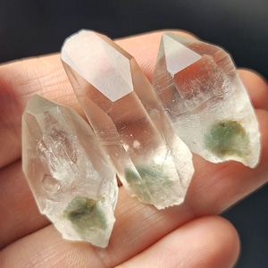 May include: Three clear quartz crystal points with visible inclusions of green minerals. The crystals are held in a hand, showcasing their natural, rough-hewn shapes and translucent quality. The crystals vary in size and have a slightly frosted appearance.