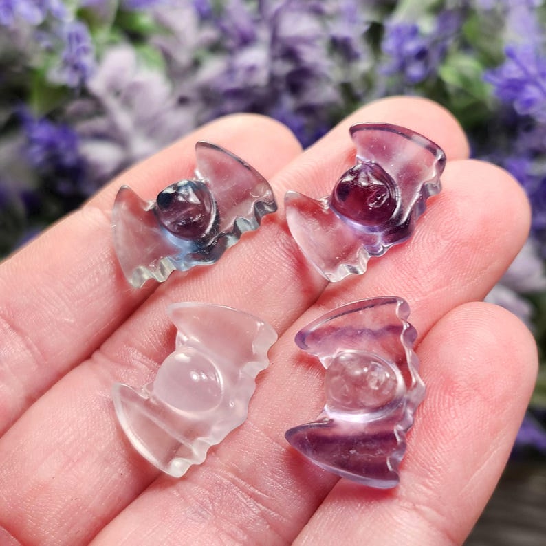 May include: Four small, translucent crystal carvings in the shape of a bat. The carvings are in shades of purple, blue, and clear. The carvings are held in a hand, with a blurred floral background.