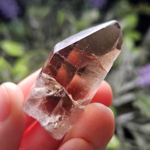 Puede incluir: Un cristal de cuarzo ahumado claro y facetado, de color marr&oacute;n. El cristal se sostiene en una mano, con un fondo borroso verde y morado. Las facetas del cristal capturan la luz, mostrando su belleza natural.