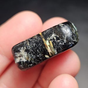 May include: A polished, rectangular gemstone with a black base color and iridescent inclusions of gold, green, and white. The stone has a smooth, rounded shape and a glossy finish. This unique mineral specimen is ideal for collectors.