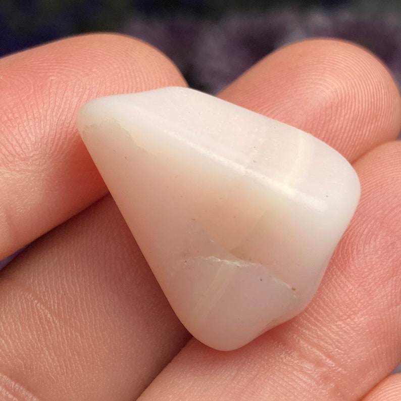 May include: A smooth, polished, opaque white stone with a triangular shape and subtle, faint horizontal banding. The surface of the stone features a small, thin, natural fissure line across the lower section, showcasing a milky, off-white colour.