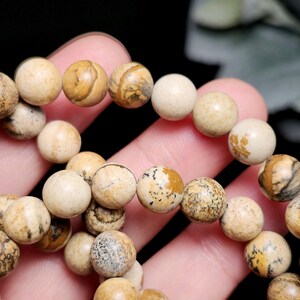 May include: A close-up of a bracelet made of round, beige-colored beads. The beads have unique patterns of brown and tan, with some appearing solid. The bracelet is held in a hand, showcasing the natural stone texture and color variations.