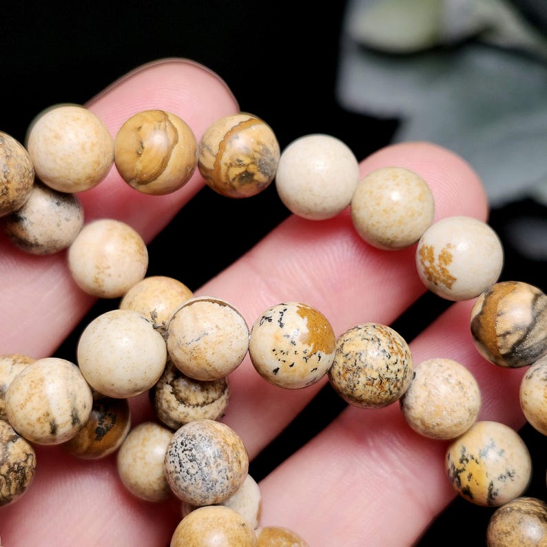 May include: A close-up of a bracelet made of round, beige-colored beads. The beads have unique patterns of brown and tan, with some appearing solid. The bracelet is held in a hand, showcasing the natural stone texture and color variations.