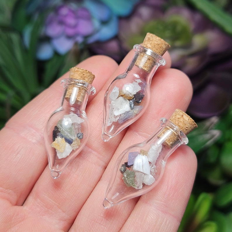 Puede incluir: Tres peque&ntilde;as botellas de vidrio transparente con tapones de corcho, cada una llena de una variedad de piedras preciosas de colores. Las botellas se sostienen en una mano, con un fondo borroso de follaje verde y morado.