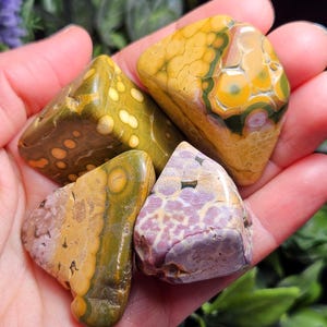 May include: Four polished gemstones in shades of yellow, green, and purple are held in a hand. The stones have unique patterns and textures, with spots and swirls. The gemstones are likely used for decorative or metaphysical purposes.
