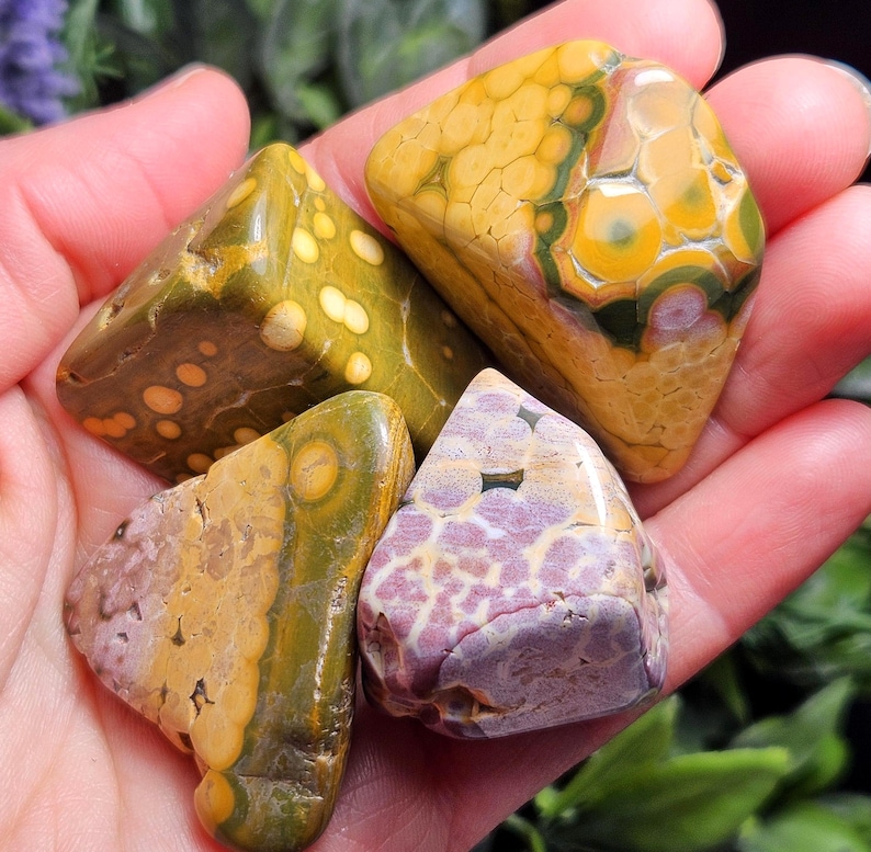 May include: Four polished gemstones in shades of yellow, green, and purple are held in a hand. The stones have unique patterns and textures, with spots and swirls. The gemstones are likely used for decorative or metaphysical purposes.