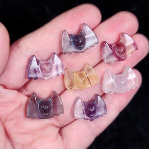 May include: A collection of small, translucent bat figurines in various colors, including purple, yellow, and clear. The bat figurines are held in a hand, showcasing their intricate details and unique shapes. The bats are likely made of a crystal or glass material.