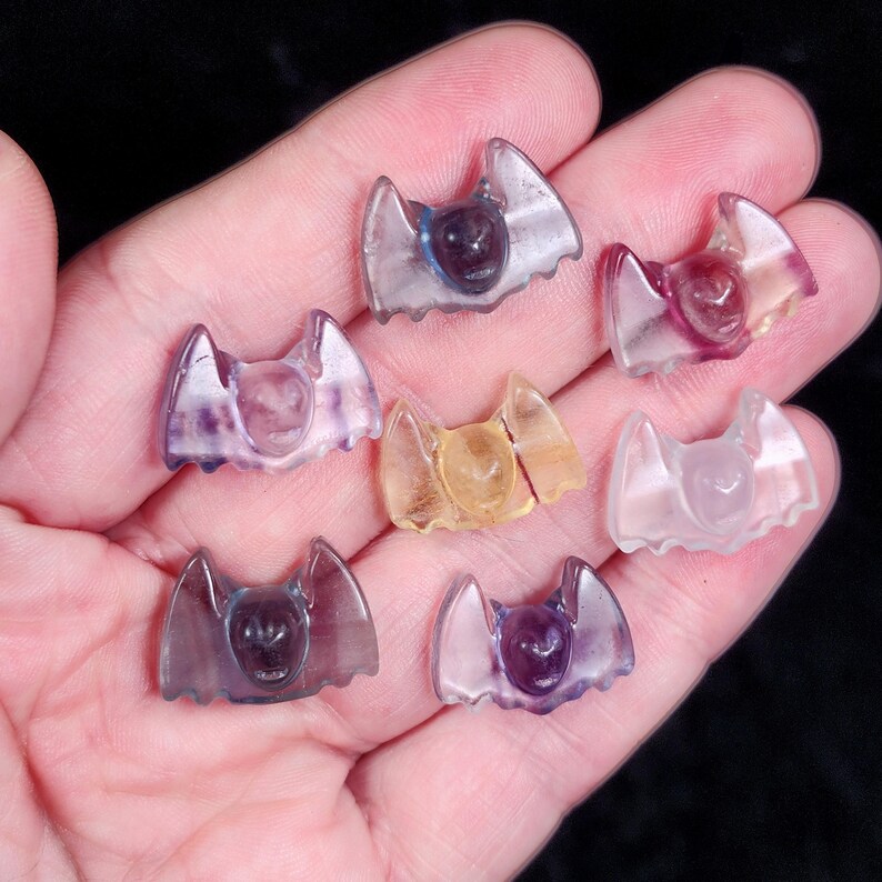 May include: A collection of small, translucent bat figurines in various colors, including purple, yellow, and clear. The bat figurines are held in a hand, showcasing their intricate details and unique shapes. The bats are likely made of a crystal or glass material.