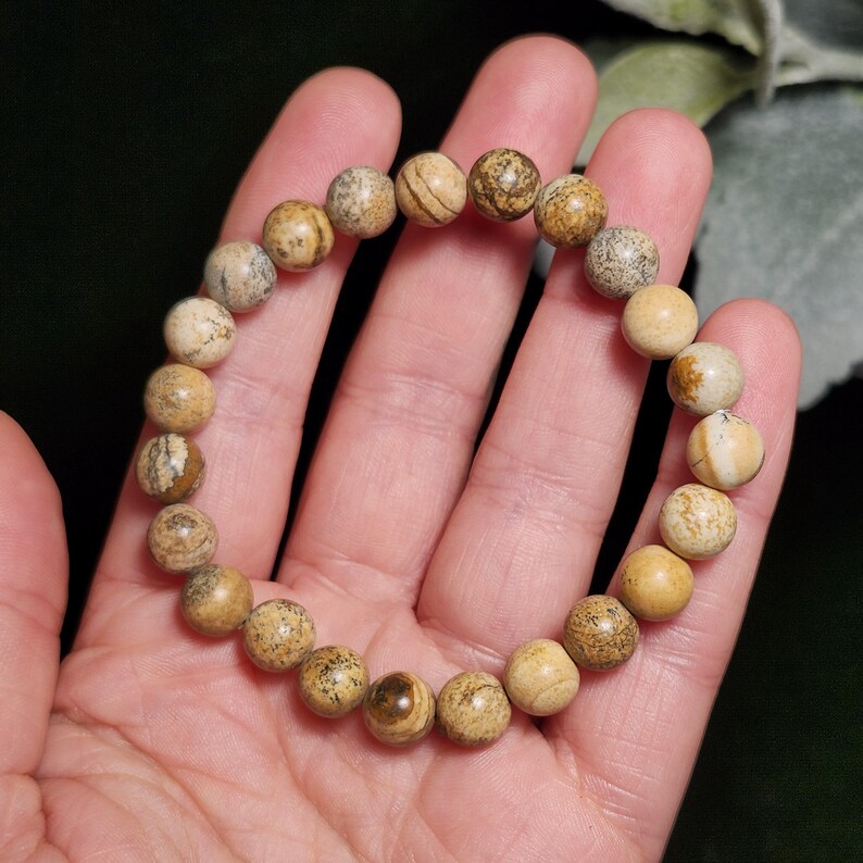May include: A beaded bracelet featuring round, earth-toned jasper stones. The bracelet is composed of multiple beads in shades of tan, beige, and brown, with unique patterns and textures on each stone. The bracelet is held in a hand, showcasing its natural beauty.