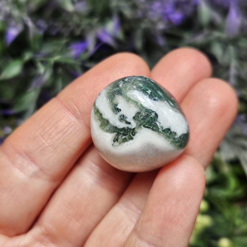 May include: A polished, rounded stone featuring a smooth surface with swirling patterns of opaque white and deep forest green. The stone has a glossy, reflective finish and a compact, pebble-like shape that fits within the palm of a hand.