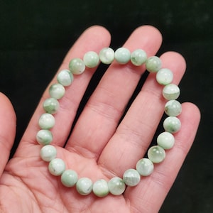 Peut inclure: Un bracelet de perles vert clair. Le bracelet est compos&eacute; de perles rondes et polies, avec des tons vert p&acirc;le et blanc. Les perles sont enfil&eacute;es pour former un cercle, adapt&eacute; au poignet. Le bracelet est pr&eacute;sent&eacute; sur un fond sombre. Chaque perle mesure environ 0,8 cm de diam&egrave;tre.