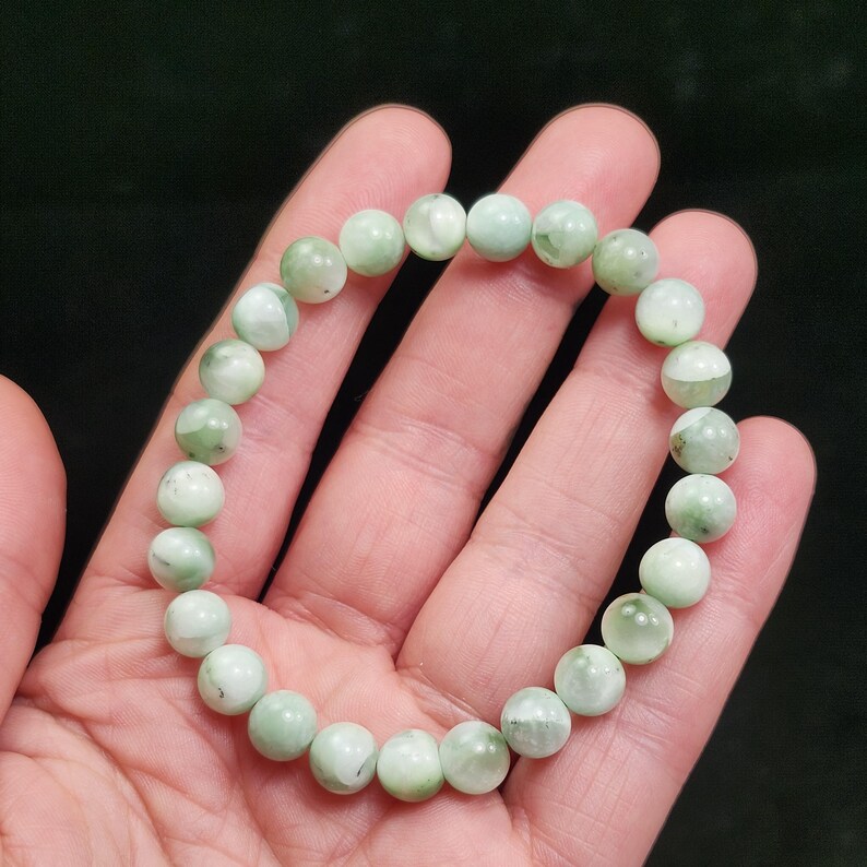 Peut inclure: Un bracelet de perles vert clair. Le bracelet est compos&eacute; de perles rondes et polies, avec des tons vert p&acirc;le et blanc. Les perles sont enfil&eacute;es pour former un cercle, adapt&eacute; au poignet. Le bracelet est pr&eacute;sent&eacute; sur un fond sombre. Chaque perle mesure environ 0,8 cm de diam&egrave;tre.