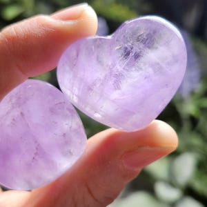 Puede incluir: Dos cristales de amatista en forma de coraz&oacute;n, uno m&aacute;s grande que el otro, con un tono p&uacute;rpura claro y una apariencia transl&uacute;cida.