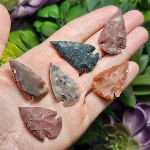 May include: Six arrowheads of various colors and patterns, including black, gray, brown, and red, are displayed on a hand. The arrowheads are made of stone and have a pointed tip.