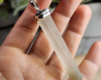 May include: A clear, rectangular pendant necklace with a silver chain. The pendant is a pale, translucent color with a silver cap. The necklace is held in a hand, showcasing the pendant's shape and clarity.