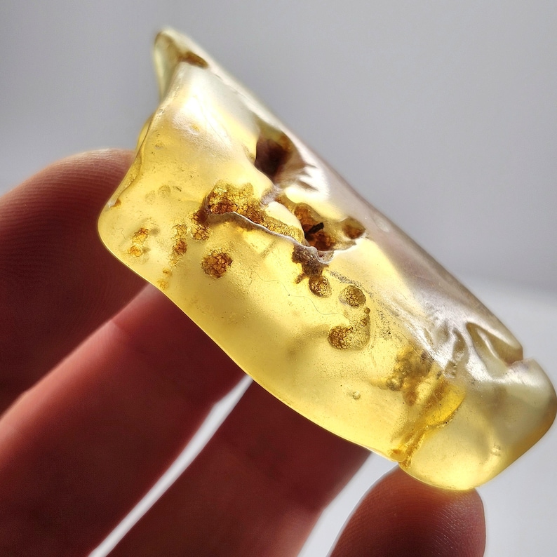 May include: A translucent, polished piece of golden-yellow amber with visible internal organic inclusions and natural fissures. The irregular, smooth-surfaced stone displays varying shades of honey and light yellow, catching the light.