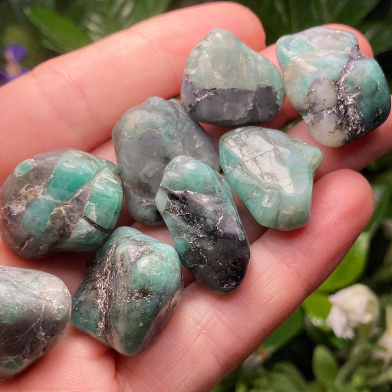 May include: A selection of raw emerald stones, displaying a range of green and black hues. The stones have irregular shapes and appear to be polished, with natural inclusions visible. The emeralds are held in a hand, showcasing their natural beauty.