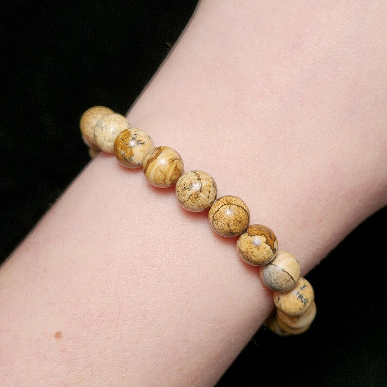 May include: A beaded bracelet featuring round, natural-toned jasper stones. The bracelet's beads display a mix of tan, brown, and cream colors, with unique patterns on each stone. The bracelet is strung together, creating a simple yet elegant accessory.