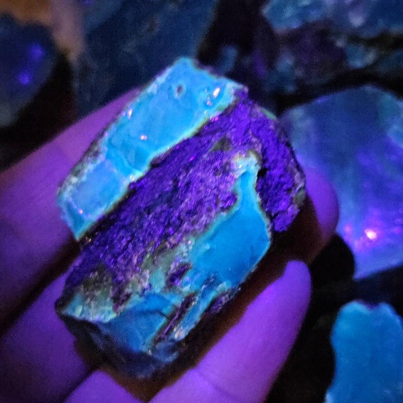 May include: A large, rough, blue-green mineral specimen with a purple hue under UV light. The mineral has a layered, striated texture.