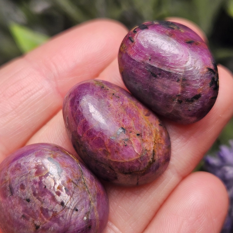 May include: Three polished, oval-shaped gemstones in varying shades of deep pink and purple. The stones have a smooth, glossy surface with natural inclusions and variations in color. The gemstones are held in a hand.