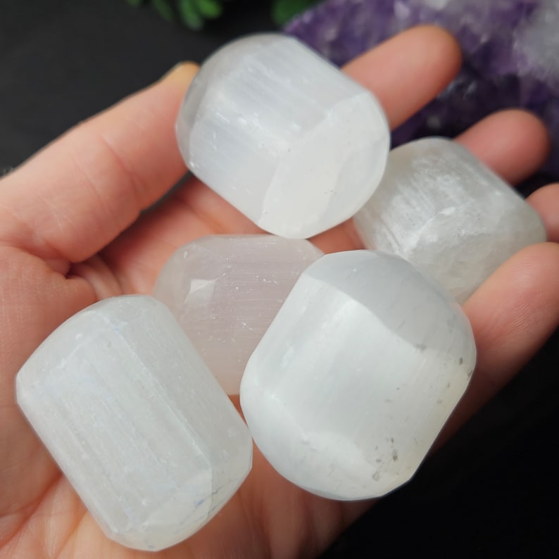 May include: Five white selenite tumbled stones, each with a smooth, polished surface and a slightly rectangular shape. The stones are arranged in a hand, with the largest stone in the centre.