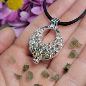 May include: A silver filigree pendant with a crescent moon shape, hanging from a black cord. The pendant is filled with small, green, translucent crystals.