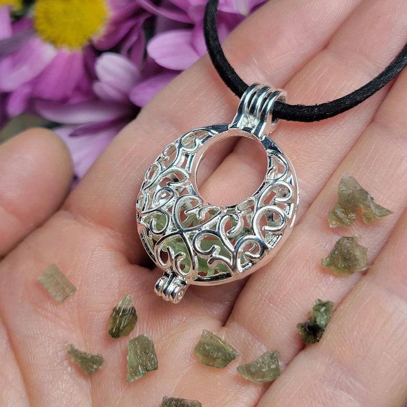May include: A silver filigree pendant with a crescent moon shape, hanging from a black cord. The pendant is filled with small, green, translucent crystals.