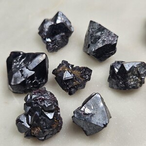 Puede incluir: Una colecci&oacute;n de cristales oscuros y facetados con brillo met&aacute;lico. Los cristales var&iacute;an en tama&ntilde;o y forma, con bordes afilados y un color negro intenso. La disposici&oacute;n sugiere una exhibici&oacute;n de espec&iacute;menes minerales en bruto, posiblemente para coleccionistas o decoraci&oacute;n.