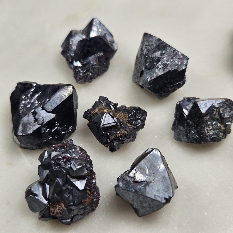 Puede incluir: Una colecci&oacute;n de cristales oscuros y facetados con brillo met&aacute;lico. Los cristales var&iacute;an en tama&ntilde;o y forma, con bordes afilados y un color negro intenso. La disposici&oacute;n sugiere una exhibici&oacute;n de espec&iacute;menes minerales en bruto, posiblemente para coleccionistas o decoraci&oacute;n.