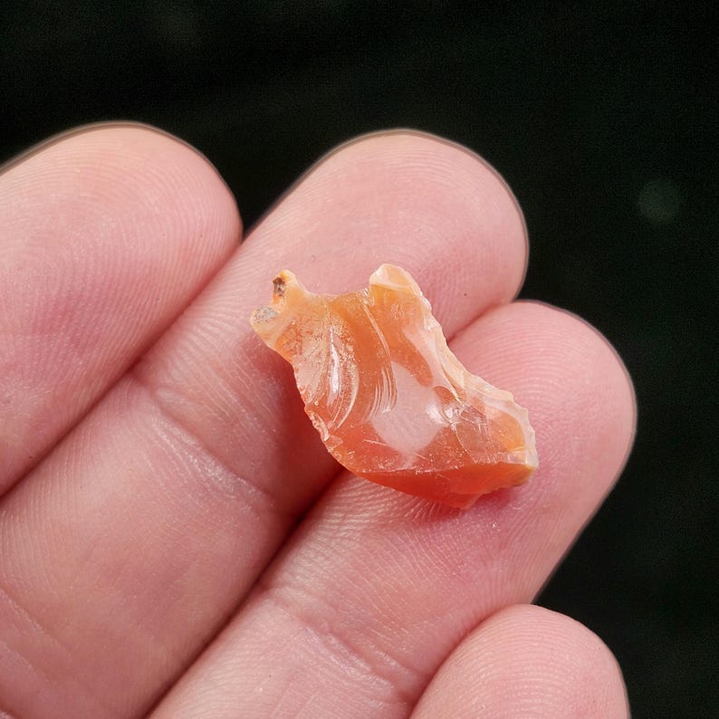 May include: A small, translucent, orange-colored gemstone with a rough, irregular shape. The stone is held in a person's hand against a dark background. The gemstone has a polished, glassy appearance.
