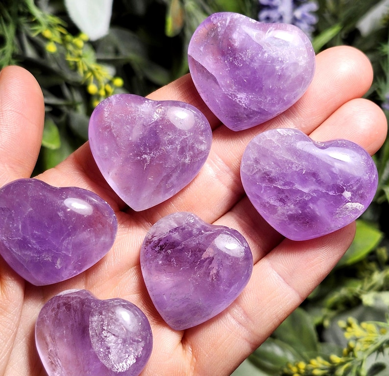 May include: Six heart-shaped amethyst crystals in various sizes, displayed on a hand. The crystals are a deep purple color and have a smooth, polished finish.