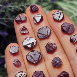 K&ouml;nnte beinhalten: Eine Sammlung von 15 tiefvioletten, facettierten Rubinkristallen in verschiedenen Gr&ouml;&szlig;en und Formen. Die Kristalle sind auf einer Hand angeordnet und zeigen ihre einzigartigen Facetten und ihren Glanz.